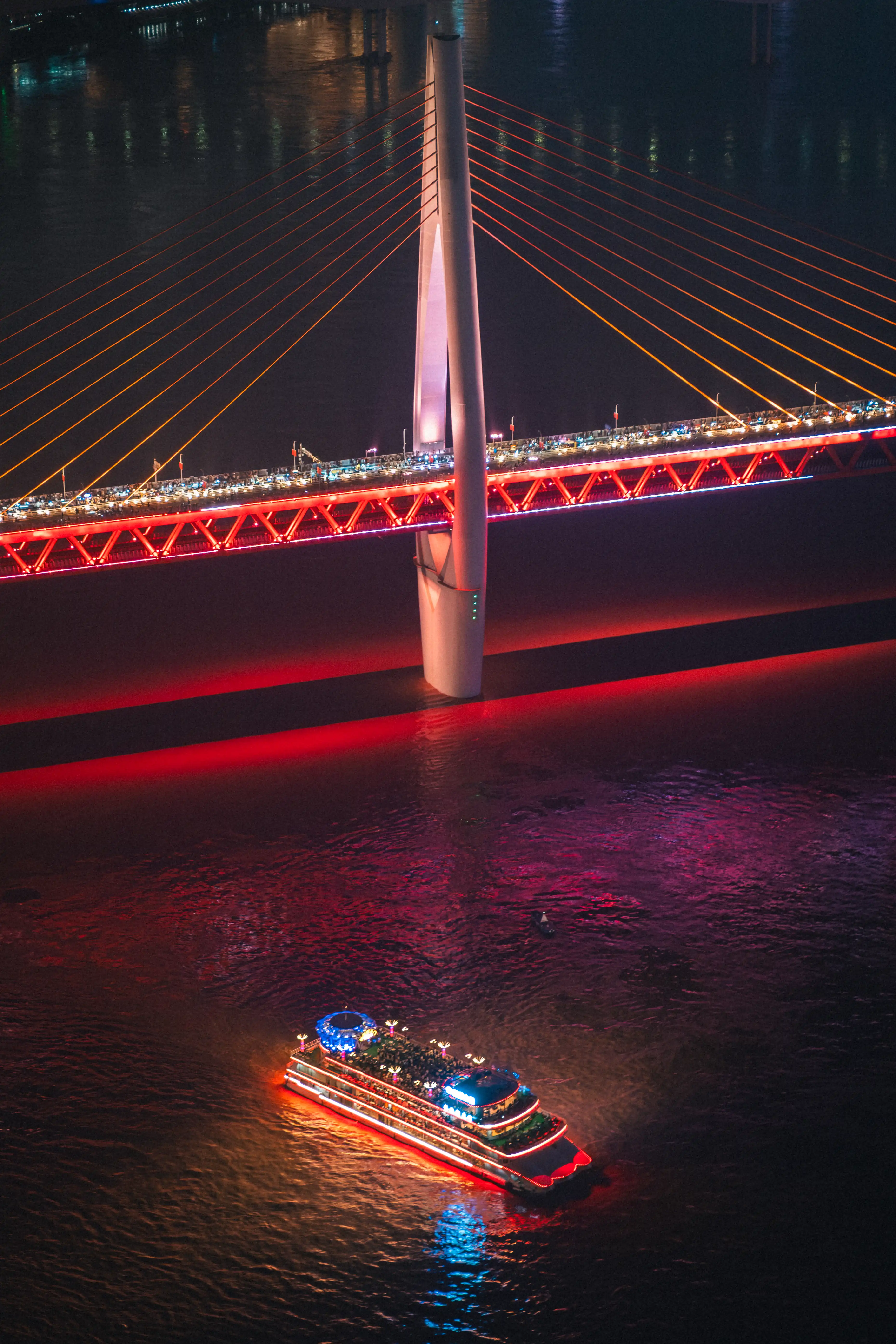 Vue plongeante nocturne sur le fleuve de Chongqing, capturant un bateau de croisière illuminé glissant sous un pont à la base du pilier principal.