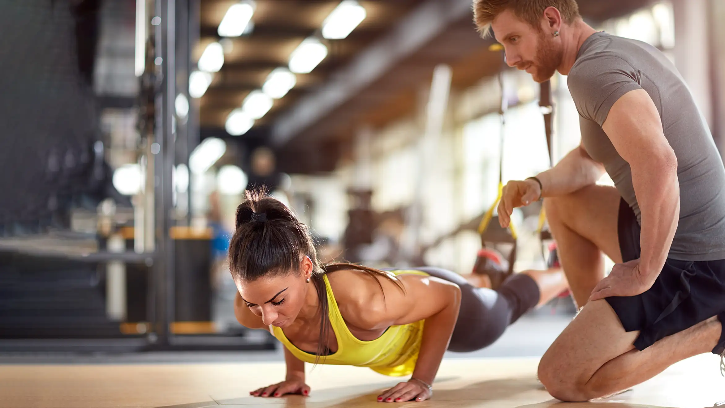 Woman doing pushups in a gym with a man kneeling by his side