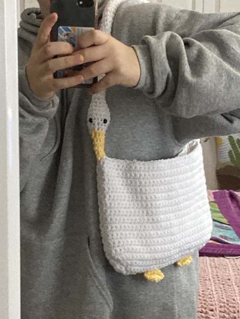 A crocheted tote that looks like a white goose. The neck is the handle of it and is attached to itself by the beak. It also has little goose feet on the bottom.