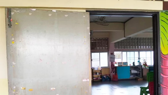 Gran panel corredero sin terminar en la entrada de un aula, con una sala de juegos al fondo.