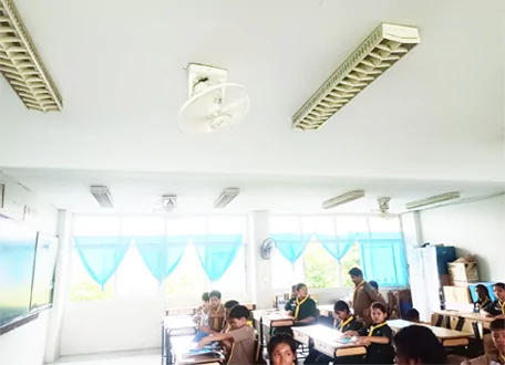 Alumnos sentados en pupitres de un aula luminosa con ventiladores de techo y luces fluorescentes.