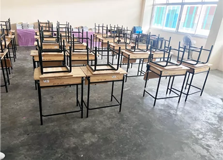 Aula en obras con pupitres de madera apilados boca abajo sobre el suelo de hormigón.