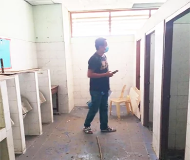 Hombre inspeccionando los aseos escolares deteriorados, con puertas de cabina ausentes y suelo dañado.