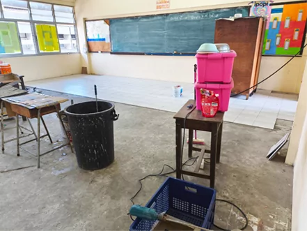 Aula durante la renovación del suelo, con pavimento retirado, un cubo de agua y herramientas para colocar baldosas.