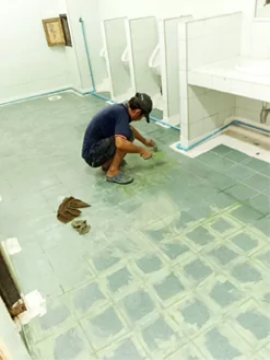Trabajador arrodillado colocando baldosas nuevas en el suelo de un aseo escolar.
