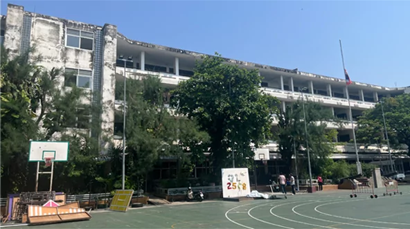 Edificio escolar de tres plantas deteriorado frente a una cancha, con árboles, asta de bandera y canasta de baloncesto antes de pintarlo.