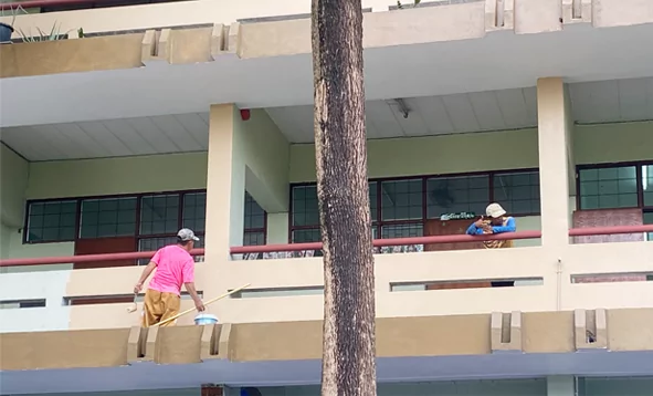 Trabajadores pintando un balcón escolar; uno, con camisa rosa, utiliza un rodillo largo.