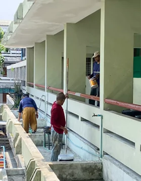 Tres trabajadores pintan y limpian un pasillo escolar; uno sostiene un bote de pintura en la planta superior.