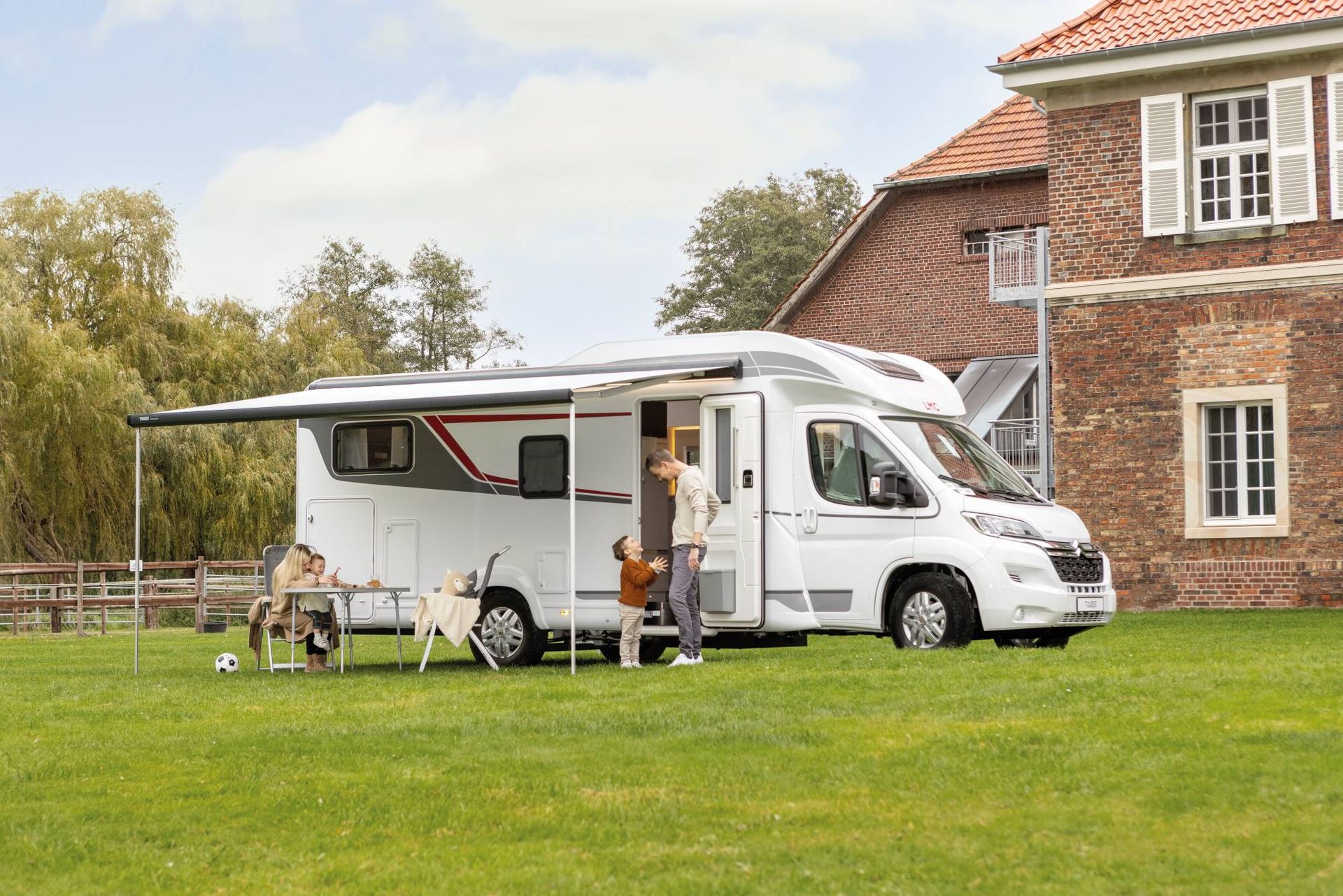 Seitlicher Blick auf das teilintegrierte Wohnmobil auf einer Wiese mit Markise und einer Familie – teilintegriertes Wohnmobil mieten Monheim