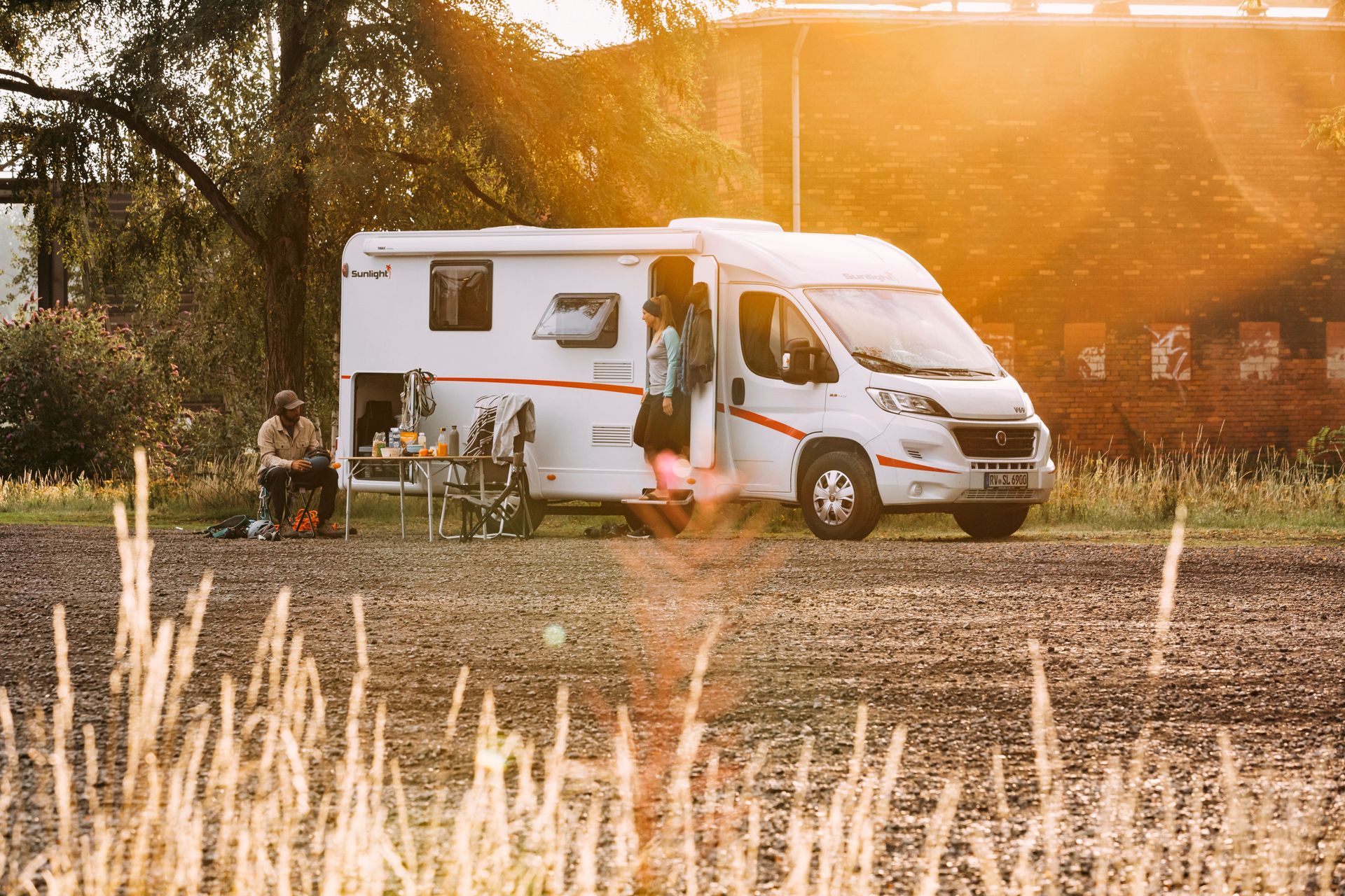 Wohnmobil auf einer Wiese mit Getreide im vorderen Bereich und indirekte Sonneneinstrahlung von vorne. Vor dem Camper ist eine Camping Garnitur aufgebaut, an der zwei Menschen sitzen.