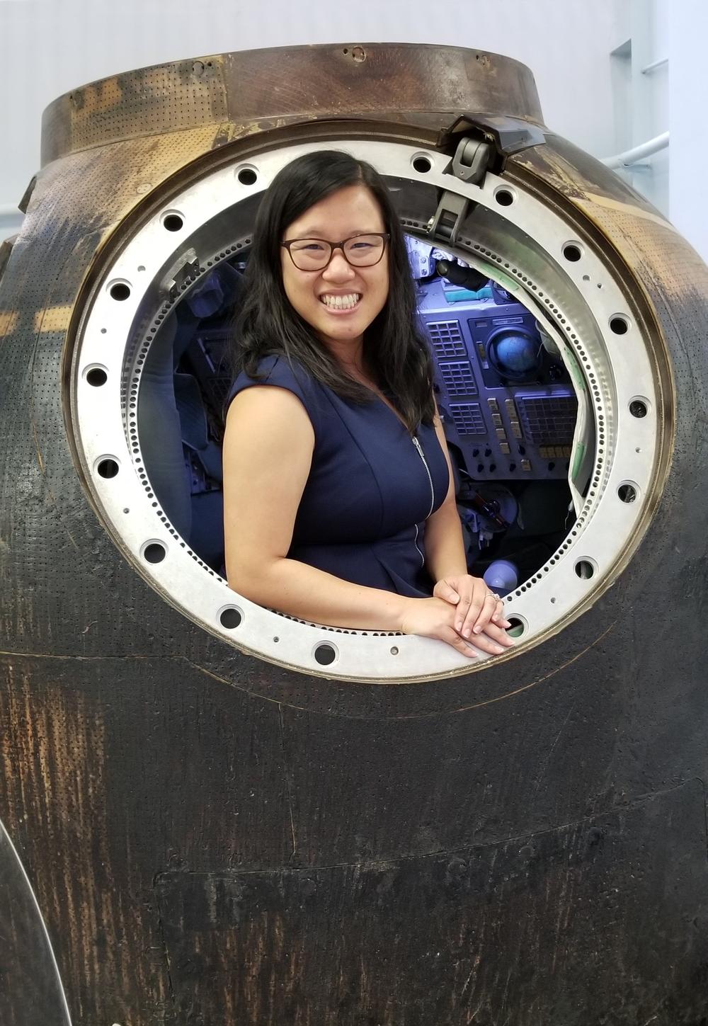 An Asian woman wearing glasses stands inside a mockup of a Russian spacecraft.