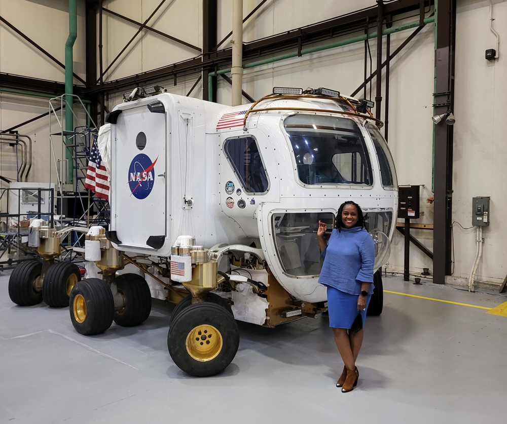 A woman stands smiling next to a NASA rover vehicle inside a large industrial facility. The woman is wearing a blue dress and brown shoes, and she is holding a small item in her hand. The American flag is visible in the background. 