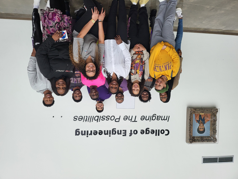 A group of students and faculty members stand in front of a wall that reads "College of Engineering" with the slogan "Imagine The Possibilities." The group is smiling and posing for the photo. A portrait of a man is displayed on the wall to the left of the group. 