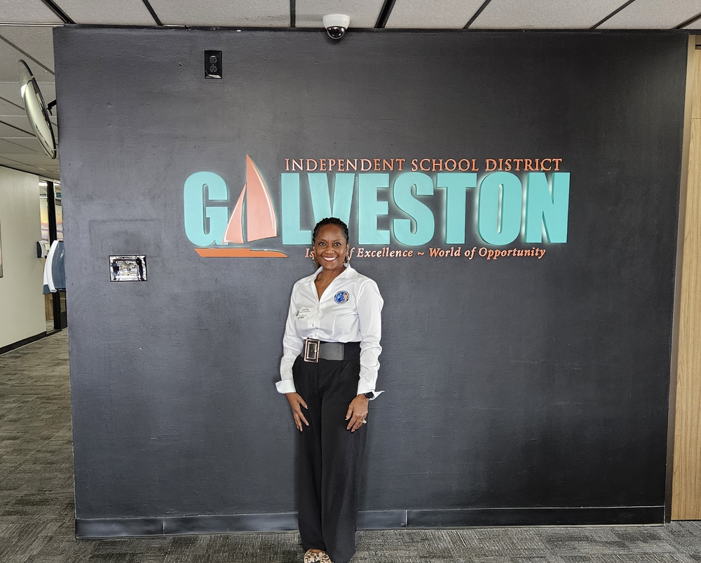A woman stands smiling in front of a wall that features the logo and text for "Galveston Independent School District" with the tagline "Island of Excellence ~ World of Opportunity." The logo includes an illustration of a sailboat. She is wearing a white blouse and black pants, standing in an indoor hallway with carpeted floors and ceiling panels.
