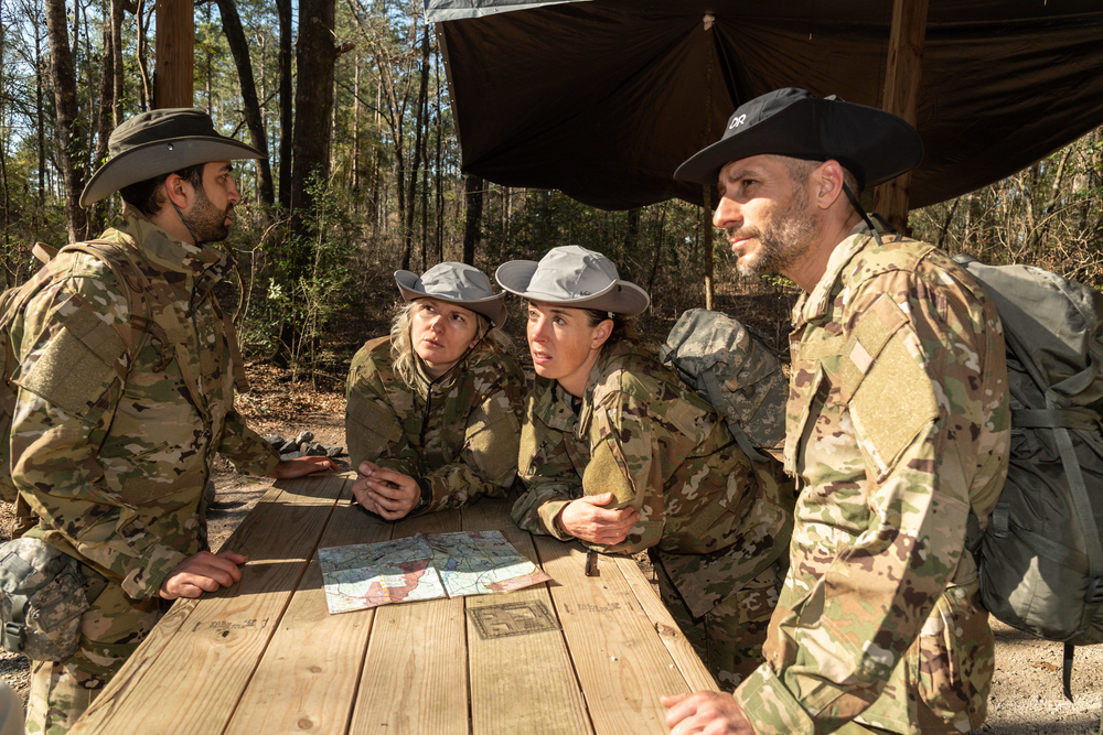 Four people around a table wear hats and a camouflaged outfit on a sunny day.