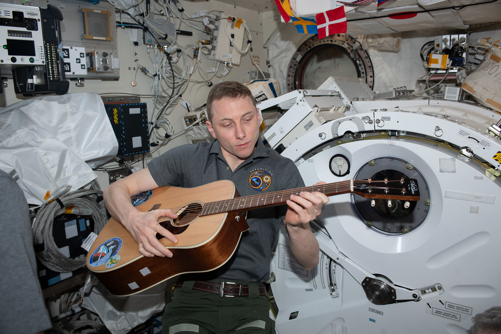 A person playing the guitar in microgravity wearing a grey polo.
