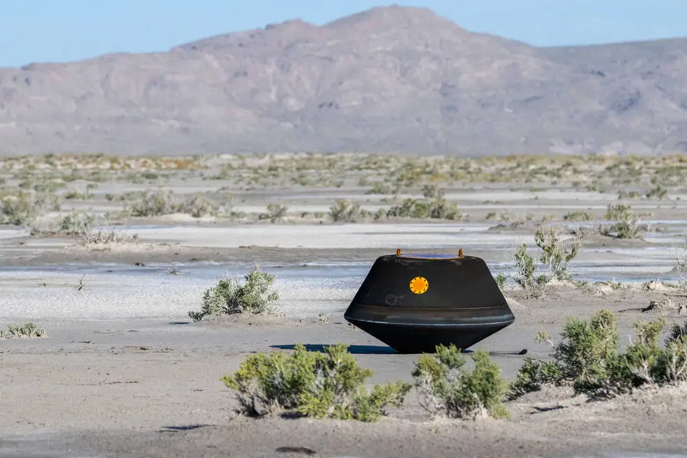 An image of a black capsule in the middle of the desert with mountains in the background.