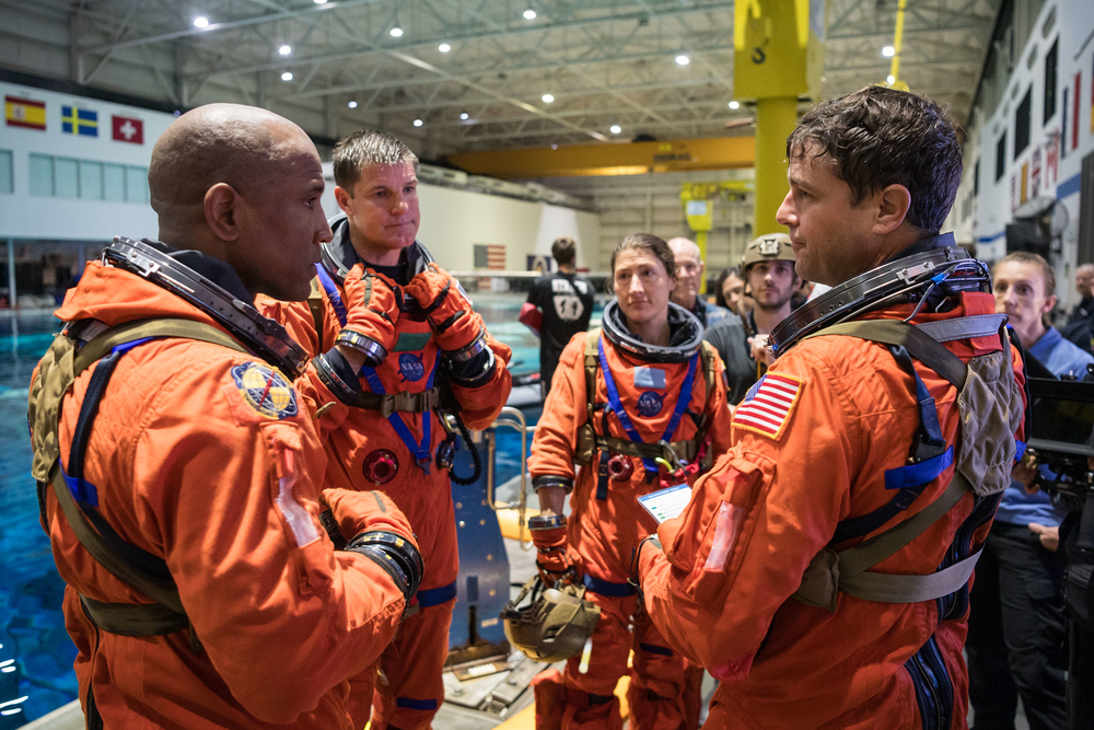 Four people in orange space suits converse in a circle beside a large pool in a facility. 