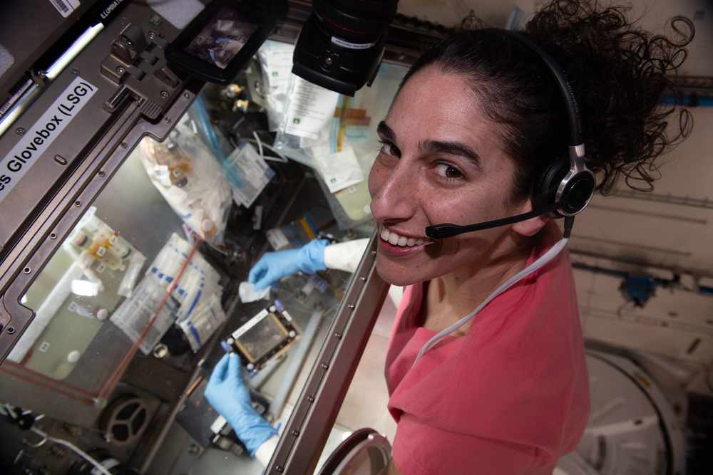 A person wearing a pink sweater looks up while performing an experiment in a microgravity laboratory.