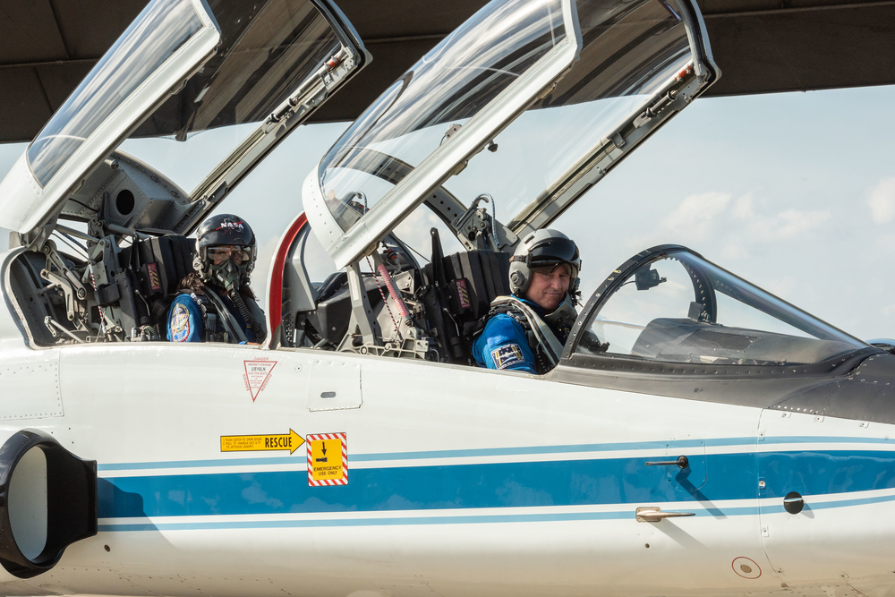 Two people wearing helmets and a blue flight suit sit inside of a jet.