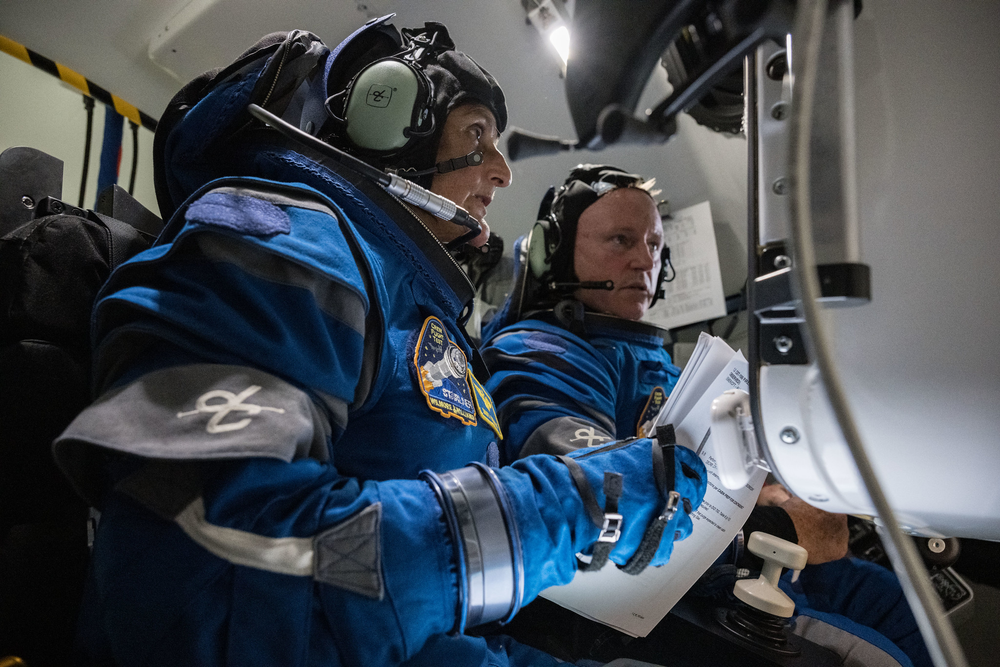 Two people wearing blue space suits sit and concentrate on instructions in a spaceflight simulator.