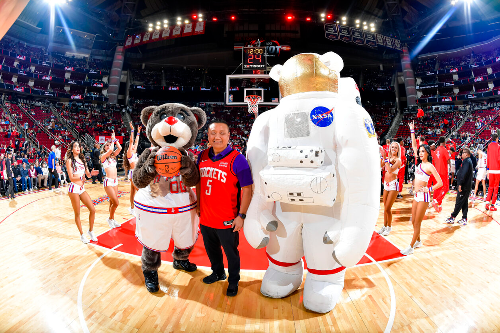 A man wearing a Houston Rockets basketball jersey stands on the court between a bear mascot and an inflatable astronaut mascot.