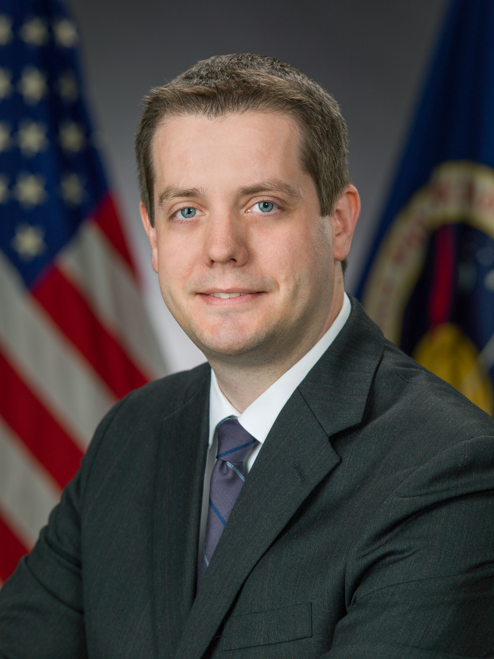 Professional portrait of a man with light brown hair, wearing a dark gray suit, white shirt, and a gray and blue patterned tie.