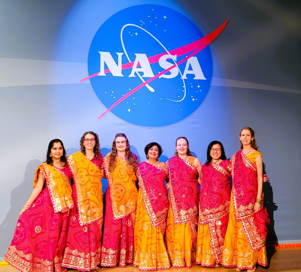 A group of seven people dressed in matched orange and red ceremonial gowns pose for a photo in front of a blue background and NASA logo.