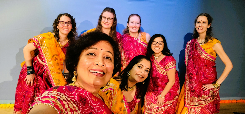 Seven people dressed in red and orange gowns take a selfie in front of a blue background.