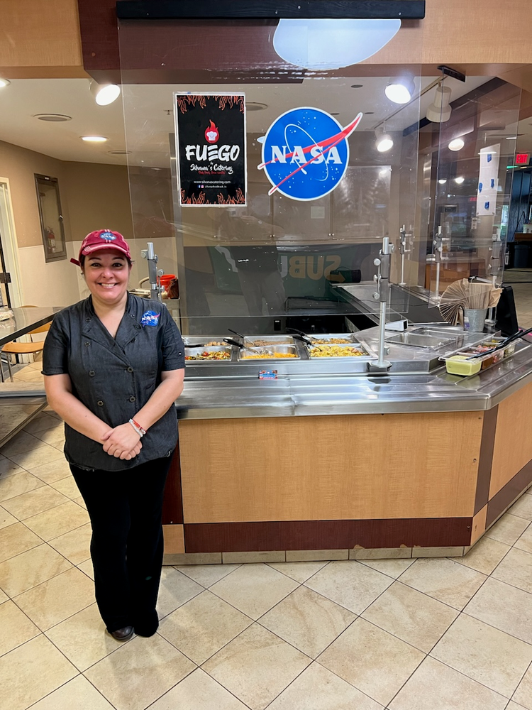 A woman in a NASA-themed uniform stands smiling in front of a cafeteria-style food service counter. Above the counter, there is a "Fuego Silvana's Catering" sign alongside a NASA logo. 