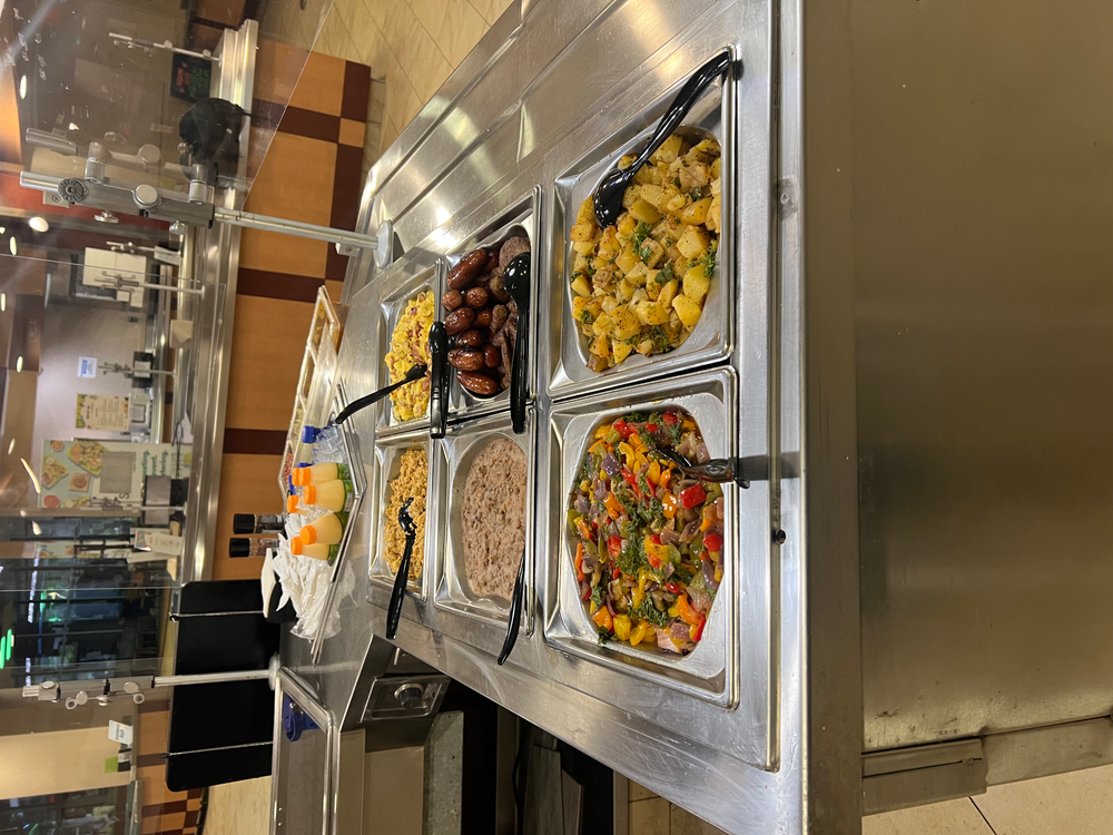 A cafeteria-style food service counter featuring a variety of dishes. The dishes include scrambled eggs, sausages, refried beans, a colorful vegetable mix, and roasted potatoes. On the counter, there are also trays of sauces, condiments, and beverages like orange juice.