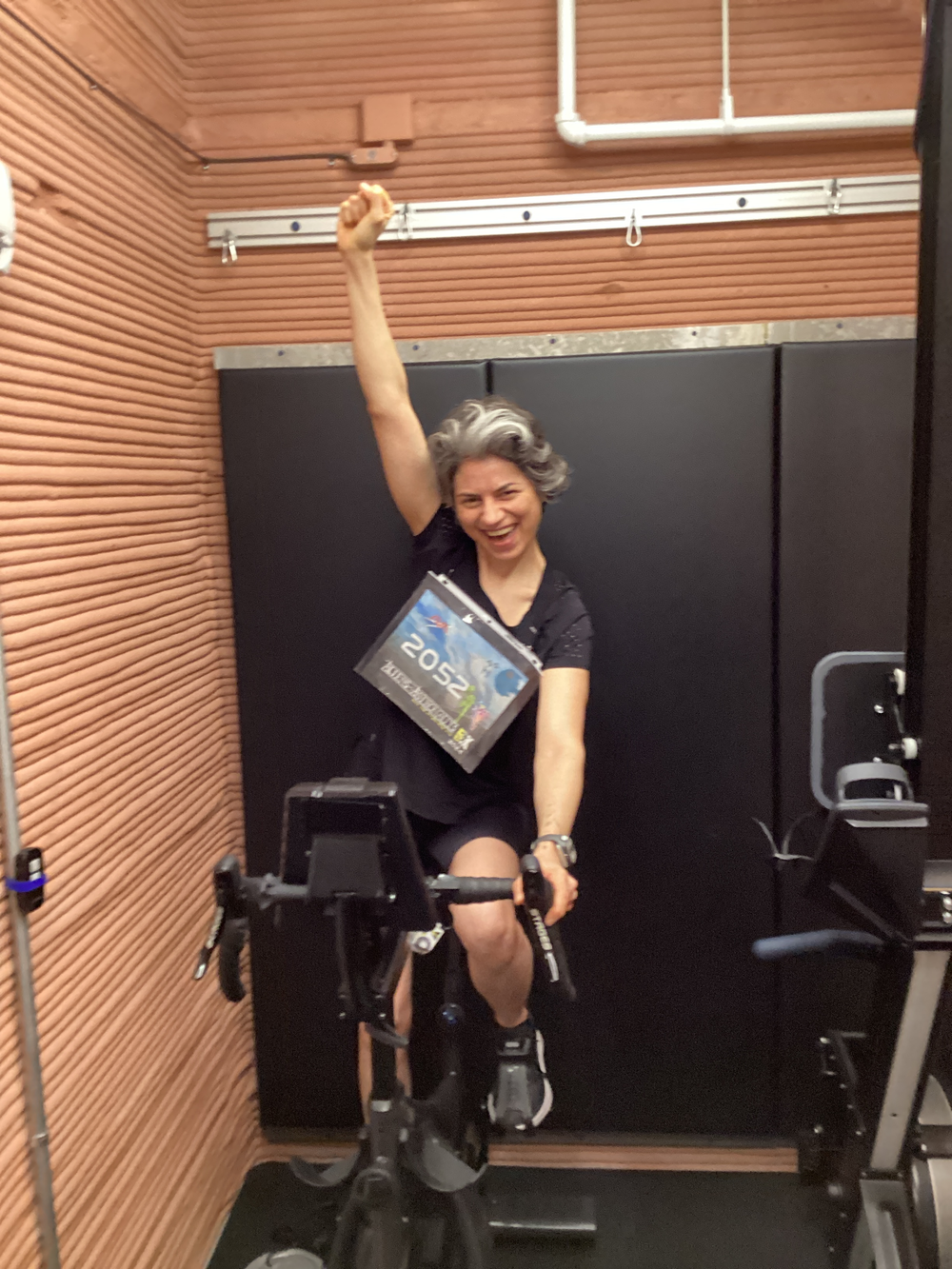 A woman with short gray hair, wearing a black t-shirt and shorts, raises her left arm in victory while sitting on a stationary bike. The setting includes wooden paneling and a black wall. 