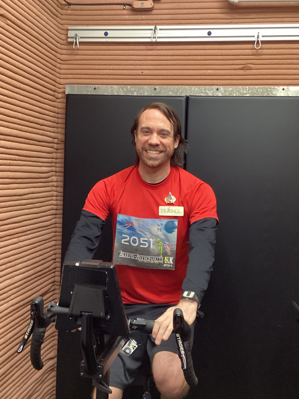 A man with short brown hair, wearing a red t-shirt and shorts, smiles while sitting on a stationary bike. The setting includes wooden paneling and a black wall.