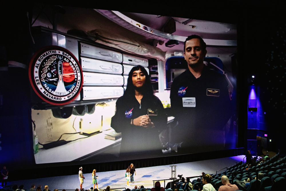 A large screen in an auditorium displays two NASA personnel in black jumpsuits with NASA patches, speaking to the audience. They stand in a spacecraft interior with equipment and a mission patch visible on the wall. Below the screen, a few people are on the stage, and the auditorium seats are partially filled with attendees watching the presentation.