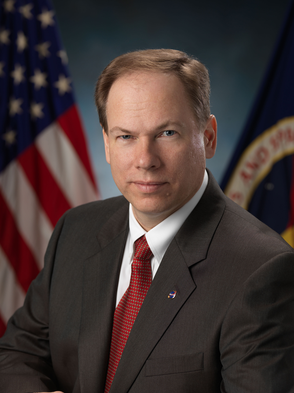 Professional portrait of a man with light brown hair, wearing a black suit, white shirt, and red tie.