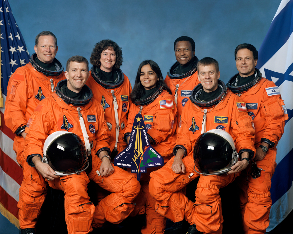 An image of seven people in orange space suits posing for a photo in front of a blue background.