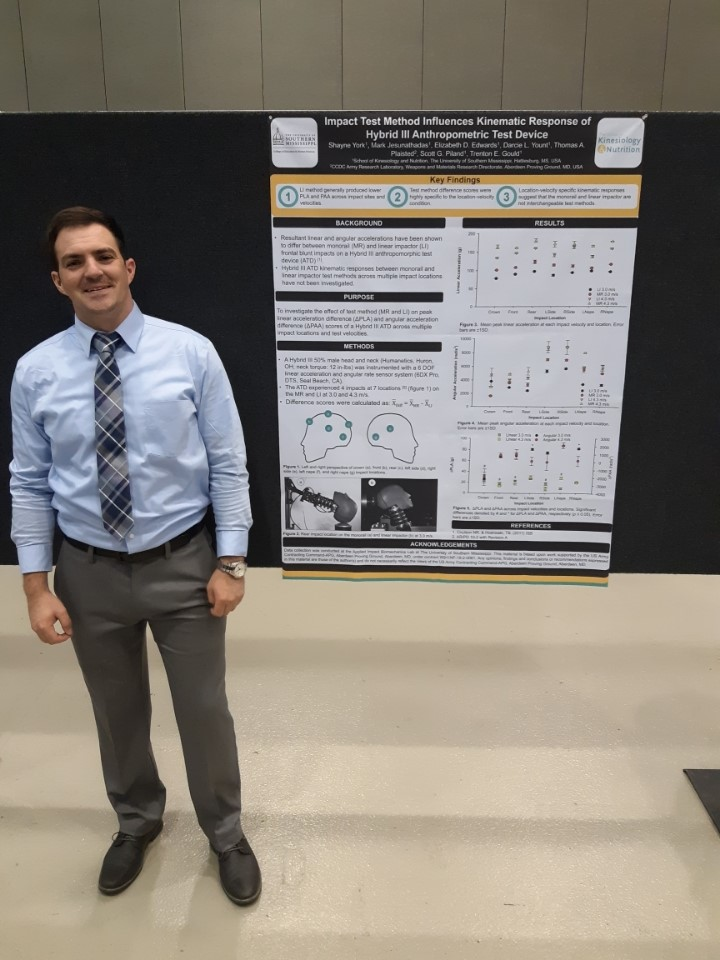 A man wearing a blue shirt and a gray-patterned tie stands in front of a poster presenting his research at a conference.