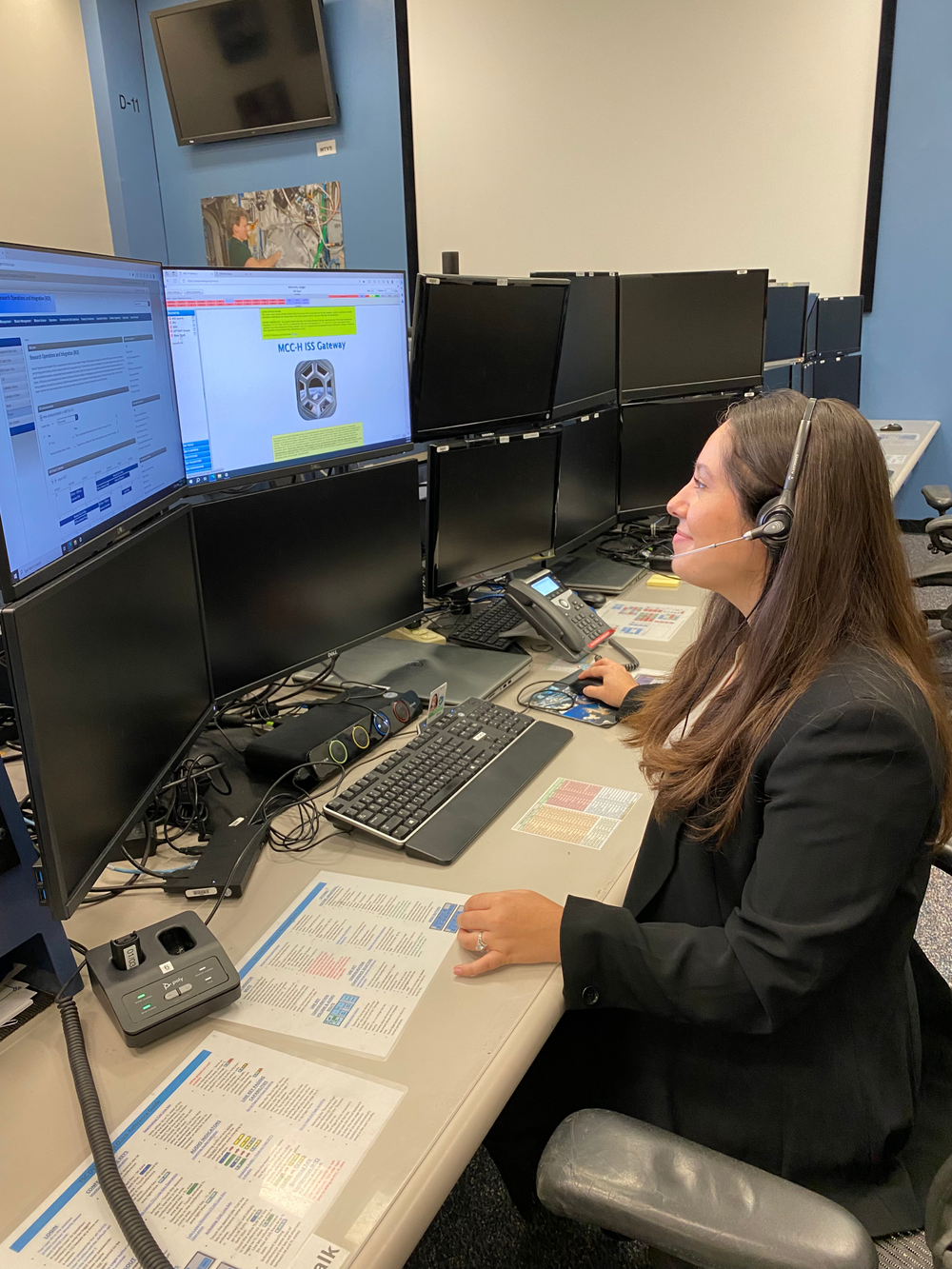 A young woman sitting at a desk looks at several large computer screens.