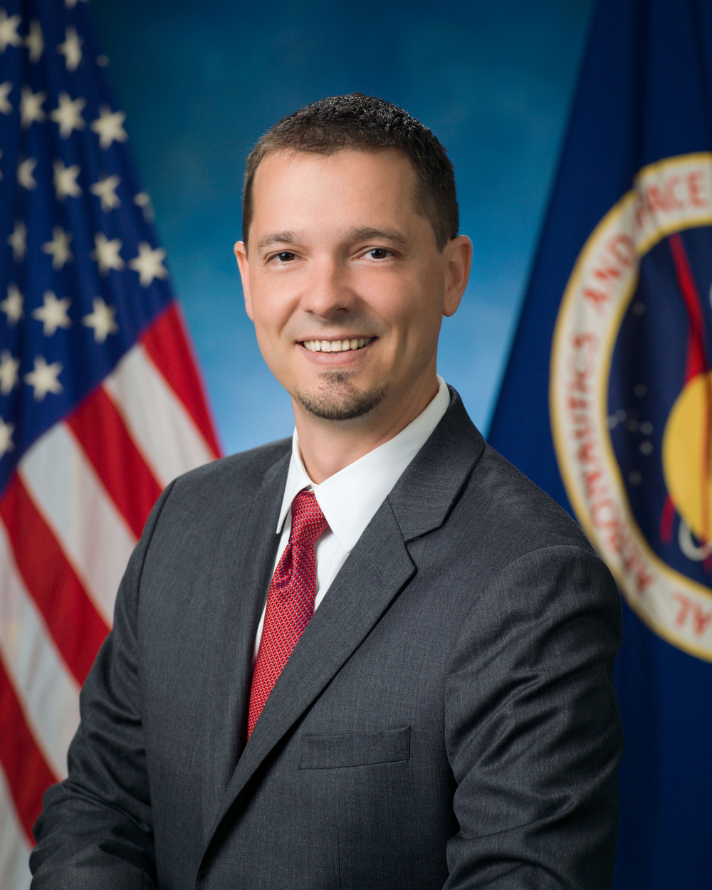 Professional portrait of a man with a goatee wearing a dark gray suit, white shirt, and red tie.