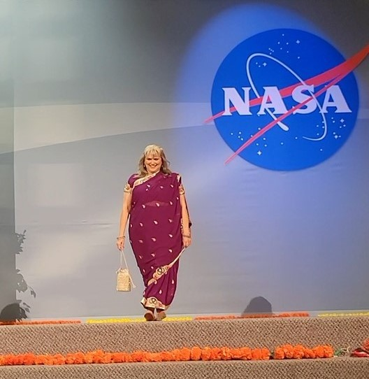 A person in a maroon and tan dress holding a tan satchel in front of a blue background and NASA logo.