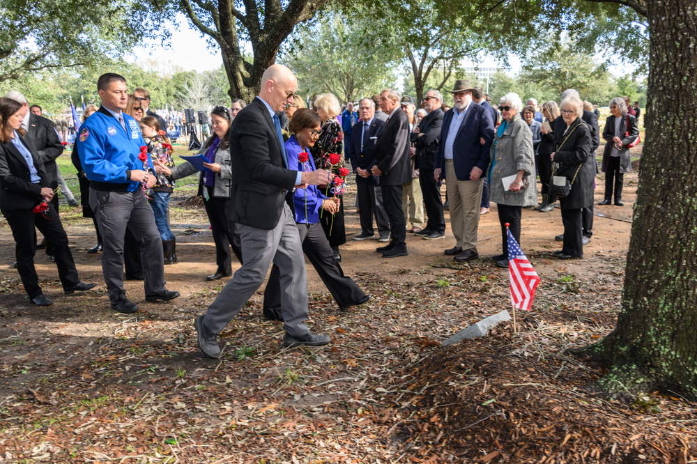 A crowd of people holding red roses walking toward a memorial under a tree with an American flag planted in the ground. 