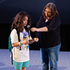 A large screen in an auditorium displays a close-up selfie of four smiling NASA personnel. They are in a spacecraft interior, with equipment visible in the background. One person is making a peace sign. The auditorium seats are partially filled with attendees watching the presentation.