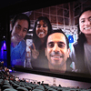 A large screen in an auditorium displays a close-up selfie of four smiling NASA personnel. They are in a spacecraft interior, with equipment visible in the background. One person is making a peace sign. The auditorium seats are partially filled with attendees watching the presentation.