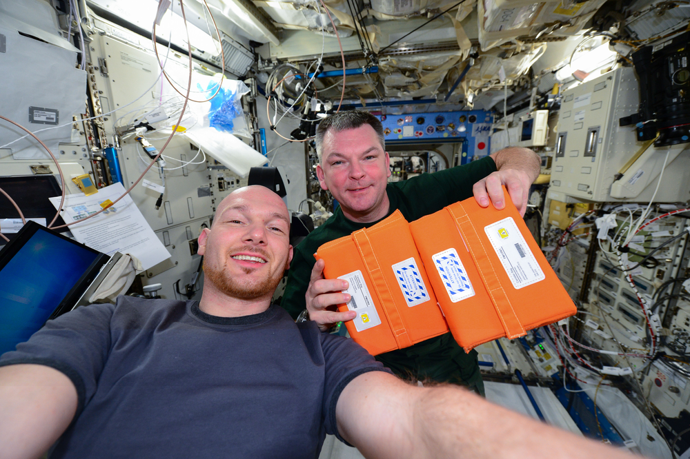 Two people in a microgravity laboratory posing for a picture. The person to the left is holding two orange rectangular boxes.