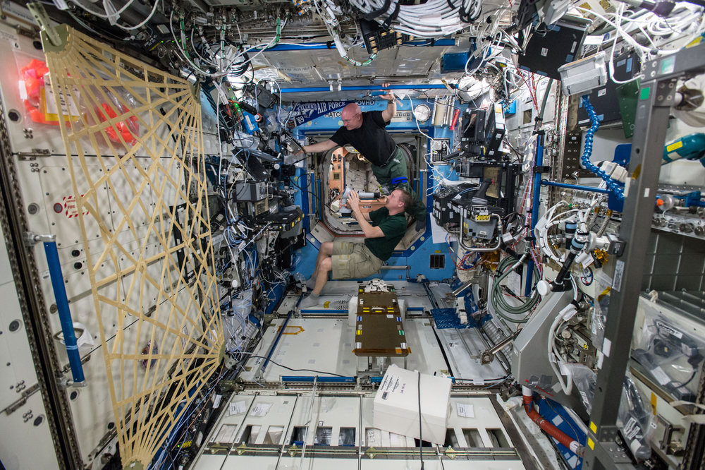 Two astronauts float inside a densely packed laboratory module on the International Space Station, surrounded by a complex array of scientific equipment, cables, and instruments. 