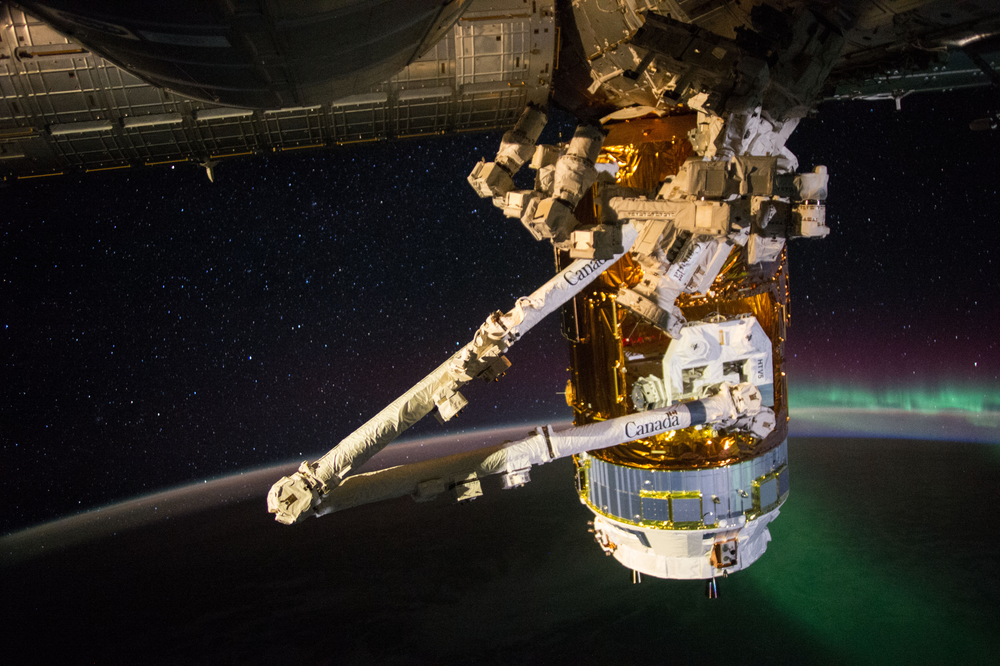 An image of an experiment and robotic arm on the side of the International Space Station with an aurora over the Earth limb visible in the background.