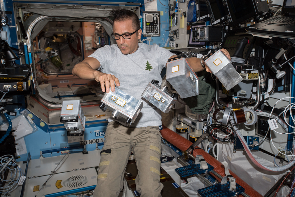 A person aboard the International Space Station wearing a grey shirt and tan pants surrounded by five canisters floating in microgravity inside a laboratory module. 