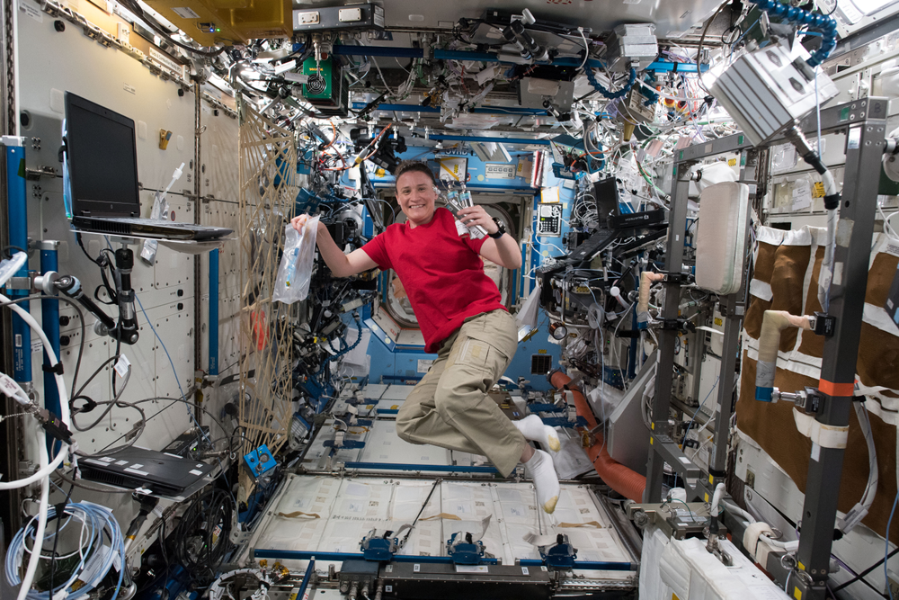 A female astronaut holds up metal tubes and plastic bags while floating on the International Space Station.