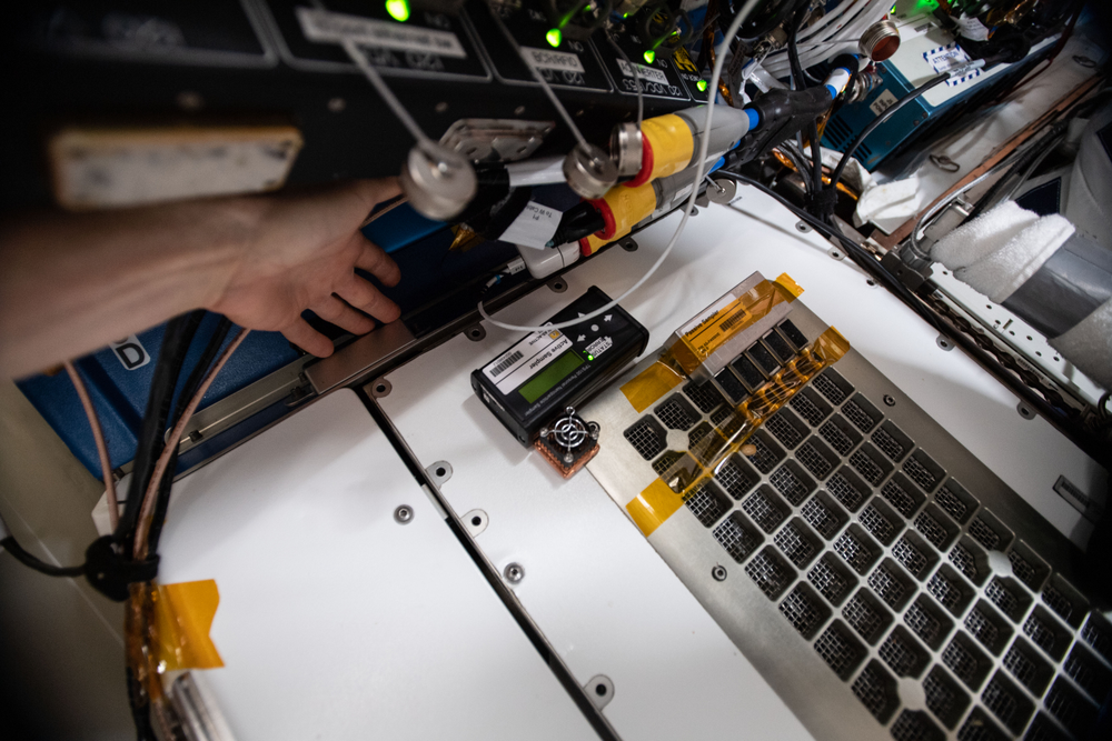 A hand reaches toward equipment on the International Space Station, featuring various cables, sensors, and a grid-like structure. 