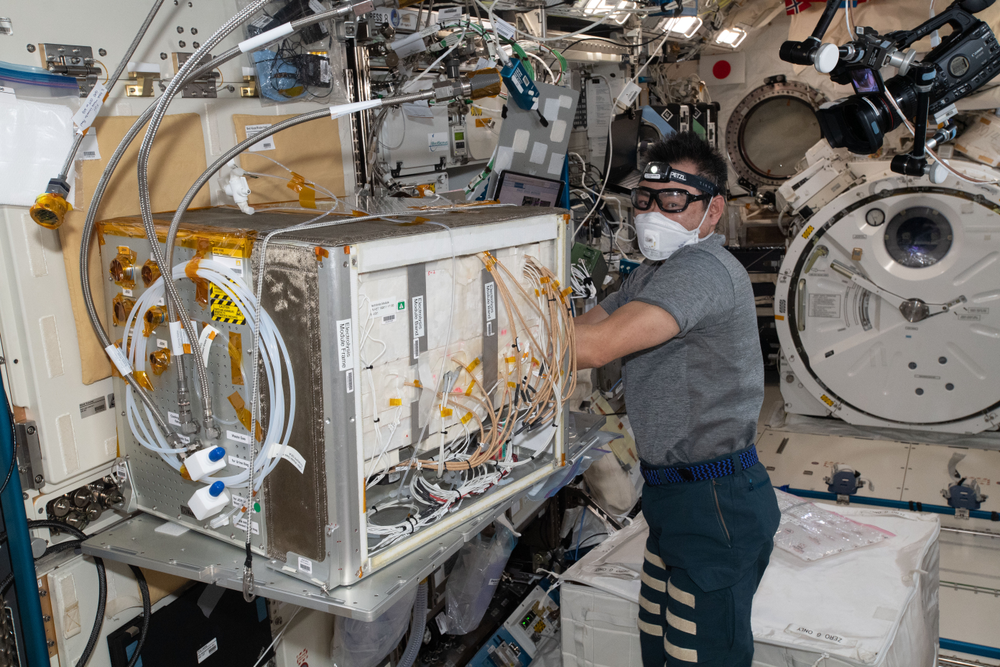 An astronaut wears safety glasses and a face mask while working with a large, box-like scientific apparatus inside the International Space Station.